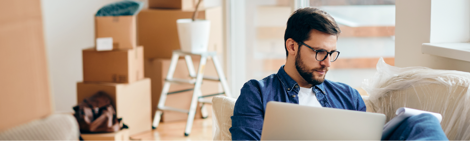 Photo of a man on a coach looking at a laptop with moving boxes and a step stool in the background.